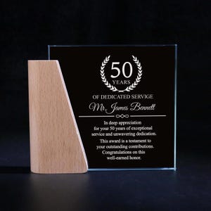 Könnte beinhalten: Eine transparente Glasauszeichnung mit schwarzem Hintergrund und Holzsockel. Die Auszeichnung trägt den Text "50 JAHRE ENGAGIERTER DIENST" und ist "Mr. James Bennett" für 50 Dienstjahre gewidmet.
