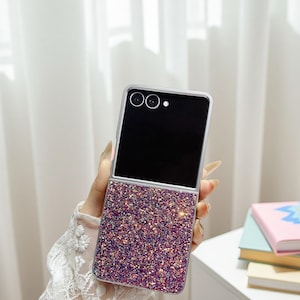 Puede incluir: Una funda plateada y negra para teléfono plegable con una mitad inferior brillante, morada y rosa. La mitad superior es negra con dos lentes de cámara. La funda se sostiene en una mano.