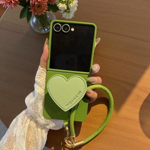 以下が含まれることがあります： ハート型のアクセントと「HAVE A NICE DAY」の文字が入った緑色の携帯電話ケース。ケースにはお揃いのグリーンのストラップとゴールドカラーの金具が付いています。携帯電話ケースは開いており、黒い画面が表示されています。