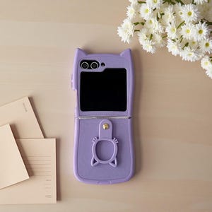 以下が含まれることがあります： 猫耳デザインのラベンダー色の携帯電話ケース。ケースは開いており、黒い画面が表示されています。金色のボタンと猫の形をしたグリップが下部にあります。白い花と文房具が背景にあります。