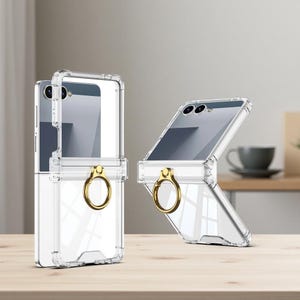 以下が含まれることがあります： ゴールドのリングホルダーが付いた透明な携帯電話ケース。ケースは折りたたみ式の携帯電話に合うように設計されています。透明なケースを通して携帯電話の画面が見えます。ケースは木製の表面にあります。