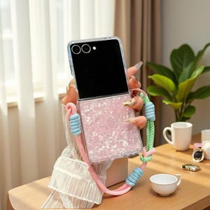 Könnte beinhalten: Eine transparente Handyhülle mit einer rosa, schillernden, strukturierten Rückseite. Die Hülle hat ein passendes rosa und blaues Schlüsselband. Der Telefonbildschirm ist schwarz. Die Hülle wird vor einem Fenster und einem Tisch mit einer Tasse und einer Pflanze gehalten.