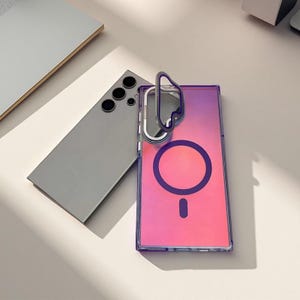 Puede incluir: Una funda transparente para teléfono con un marco morado y un fondo degradado rosa y naranja. La funda tiene un diseño circular y una pequeña barra en la parte inferior. Un teléfono gris está al lado de la funda.