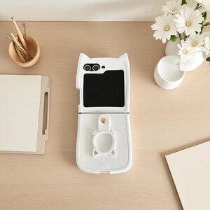 以下が含まれることがあります： 折りたたみ式携帯電話用の白い猫型携帯電話ケース。 ケースは開いており、黒い画面が表示されています。 ケースの上部には猫の顔のデザインがあり、下部には猫型の留め金が付いています。 携帯電話ケースは明るい色の木製の表面にあります。