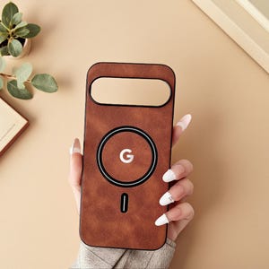 Può includere: Una custodia per telefono marrone con un ritaglio per la fotocamera e un design circolare con la lettera "G". La custodia ha una funzione di ricarica magnetica e un design minimalista. La custodia è realizzata in un materiale strutturato.