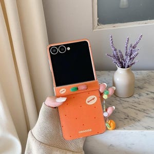 Puede incluir: Funda de teléfono naranja con diseño abatible. La funda presenta un pequeño amuleto de fruta naranja y la palabra "Orange" impresa en la parte delantera. Tiene un patrón de puntos y un pequeño diseño en forma de píldora verde y roja.