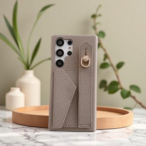 Puede incluir: Una funda de teléfono de color topo con un acabado texturizado. La funda cuenta con una ranura para tarjetas, una correa con un anillo dorado y recortes para las lentes de la cámara del teléfono. La funda se muestra en una bandeja de madera.