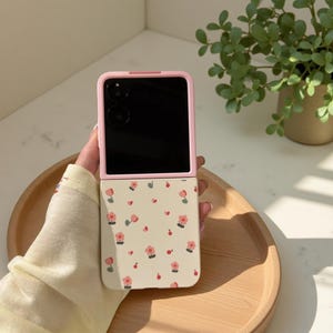 May include: A pink and cream phone case with a floral design. The case is open, showing a black screen. The floral pattern features small pink flowers and red hearts on a cream background. The case is held on a wooden tray.