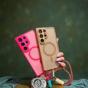 Puede incluir: Se muestran dos fundas para teléfono, una rosa y otra beige. La funda rosa tiene un anillo y un módulo de cámara rosas a juego. La funda beige tiene un anillo y un módulo de cámara beige. Una correa marrón está unida a la funda beige. Las fundas son para un smartphone.