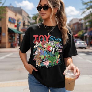 Pu&ograve; includere: T-shirt nera con una grafica colorata dei personaggi di Toy Story, tra cui Woody e Buzz Lightyear. Il testo "TOY Story" &egrave; visualizzato sopra i personaggi, con "SINCE 1995" sotto. La maglietta &egrave; indossata da una persona.