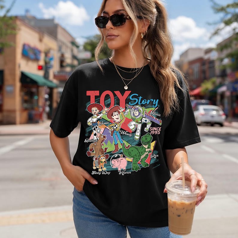 Pu&ograve; includere: T-shirt nera con una grafica colorata dei personaggi di Toy Story, tra cui Woody e Buzz Lightyear. Il testo "TOY Story" &egrave; visualizzato sopra i personaggi, con "SINCE 1995" sotto. La maglietta &egrave; indossata da una persona.