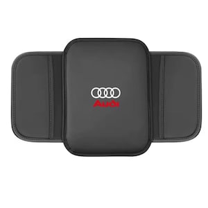 May include: A black rectangular car center console armrest cover featuring the white Audi four-ring logo and the red Audi text centered on the top surface. The cover has two side flaps with stitched edges and a smooth, matte finish throughout.