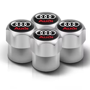 May include: Four silver tire valve stem caps with the Audi logo. Each cap has a black top with the Audi rings logo in white and the word "Audi" in red. The caps are cylindrical with a hexagonal base.