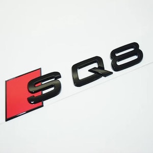 May include: Black SQ8 emblem with a red square background. The emblem is a glossy black and is likely for a vehicle. The letters are stylized and modern, with a sleek design.