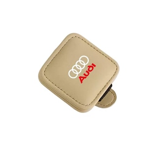 Op de afbeelding: Een vierkante, beige autostoel-kieropvuller van kunstleer met afgeronde hoeken en zichtbare stiksels langs de randen. Het Audi-logo is in wit en rood op het oppervlak gedrukt, met de vier in elkaar grijpende ringen en de merknaam.