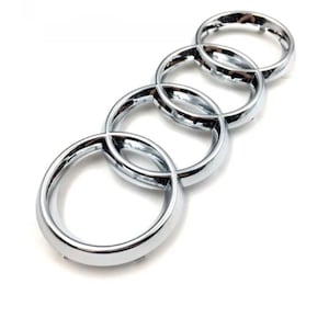 May include: A chrome-finished emblem featuring four interlocking rings. The rings are smooth, reflective, and arranged diagonally against a white background. The emblem has a modern, minimalist design.