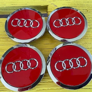 May include: Four red and silver Audi wheel center caps. Each cap features the Audi logo in silver against a red background, surrounded by a silver trim. The caps are circular and appear to be new.
