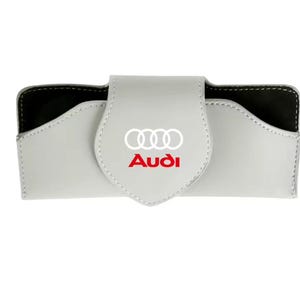 May include: A light grey rectangular glasses case with a fold-over flap closure. The front of the flap features the white Audi four-ring logo positioned above the red Audi brand name text. The interior of the case is lined with black material.