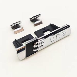 Peut inclure: Un emblème S line chromé et noir avec deux petits clips métalliques. L'emblème porte le texte "line" et la lettre "S" dans une police stylisée. L'emblème est probablement pour une voiture.