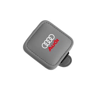 Op de afbeelding: Een vierkant, grijs, leerachtig accessoire met het witte Audi-logo van vier in elkaar grijpende ringen en de rode Audi-tekst in het midden. Het item heeft zichtbare stiksels langs de randen en een klein lipje dat aan één kant uitsteekt.