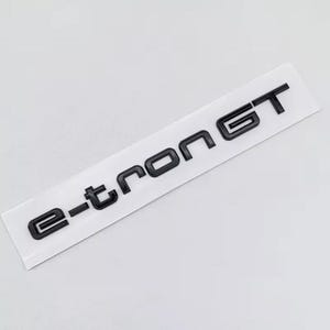 May include: Black e-tron GT emblem on a white rectangular sticker. The letters are in a bold, sans-serif font. The sticker is slightly angled against a plain white background.