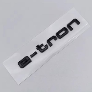 Puede incluir: Emblema e-tron negro sobre fondo blanco. Las letras en relieve y brillantes sugieren un uso automotriz. El diseño es moderno, con un ligero efecto de sombra en las letras.