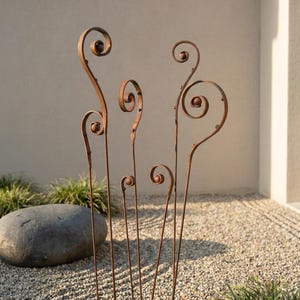 Può includere: Un set di cinque paletti decorativi in metallo per giardino con finitura rustica e usurata. Ogni paletto presenta un sottile stelo con un design a spirale arricciata nella parte superiore e un piccolo accento a sfera. I paletti sono posizionati in un letto di ghiaia.