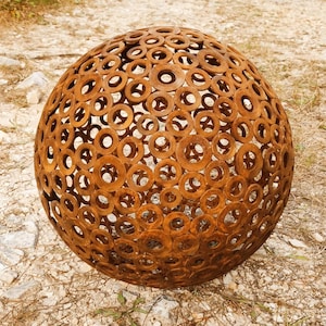 以下が含まれることがあります： 多数の錆びた金属リングで作られた、大きな球形の屋外彫刻。 リングは溶接されて中空のボールを形成し、ユニークな透かし彫りのデザインを作り出しています。 彫刻は砂利と乾燥した草の上に置かれています。