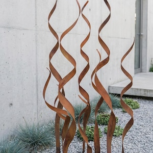 May include: A collection of outdoor metal sculptures with a swirling, ribbon-like design. The sculptures are made of a rusty brown metal and stand in a bed of grey gravel, with greenery in the background. The abstract art pieces add a modern touch to the outdoor space.