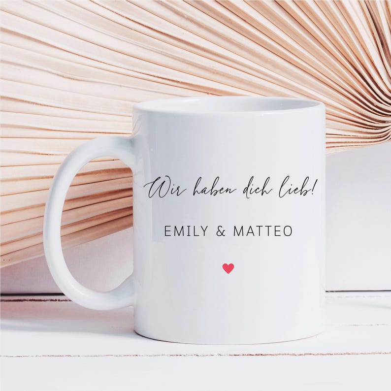 May include: White ceramic mug with a curved handle. The mug features the text "Wir haben dich lieb!" in a script font, along with the names "EMILY & MATTEO" and a small red heart. The background is a neutral color.