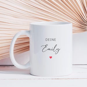 May include: White ceramic mug with a curved handle. The mug features the text "DEINE Emily" in black script, with a small red heart below the name. The background has a light brown, textured fan-like design.