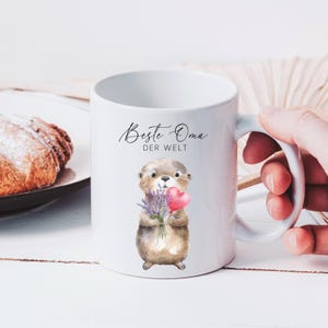 May include: White ceramic mug featuring a watercolor illustration of an otter holding lavender and a pink heart. The mug has the text "Beste Oma DER WELT" in black script. A hand is holding the mug.