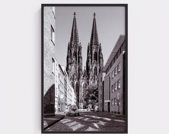 May include: Black and white framed print of a street scene in front of a cathedral. The cathedral has two tall spires. A car is parked on the cobblestone street. Buildings line the street on both sides.