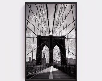 May include: Black and white framed photograph of the Brooklyn Bridge. The image captures the bridge's intricate cable structure and archways, with a wooden walkway and city skyline in the background. The photograph is a vertical composition.