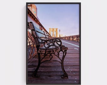 May include: A framed photograph of a bench on a wooden walkway, with the Brooklyn Bridge in the background. The bench has ornate metalwork and is made of dark wood. The sky is a soft blue.