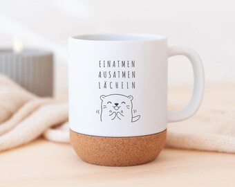 May include: White ceramic mug with a cork base. The mug features the German words "EINATMEN, AUSATMEN, LÄCHELN" and a cartoon otter illustration. The mug has a white handle and is set on a light wooden surface.