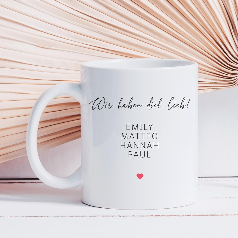 May include: White ceramic mug with a curved handle. The mug features the text "Wir haben dich lieb!" in cursive, followed by names: Emily, Matteo, Hannah, and Paul. A small red heart is printed below the names. The mug is on a white surface.