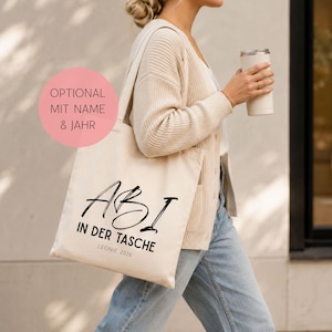 Könnte beinhalten: Eine beigefarbene Stofftasche mit dem schwarzen Aufdruck ABI IN DER TASCHE, ergänzt durch den Namen LEONIE und das Jahr 2026. Ein rosa kreisförmiges Grafikelement in der oberen linken Ecke enthält den Text OPTIONAL MIT NAME & JAHR.