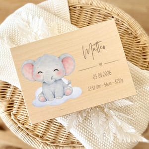 May include: A wooden plaque with the name "Matteo" in cursive script, a heart symbol, and birth details. It features a cartoon elephant sitting on a cloud, with gray and pink coloring. The plaque includes the date, time, length (56cm), and weight (3350g).