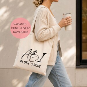 May include: A beige canvas tote bag featuring the black printed text "ABI IN DER TASCHE" in a mix of script and sans-serif fonts. A pink circular overlay in the upper left corner contains the text "VARIANTE OHNE ZUSATZ NAME/JAHR".