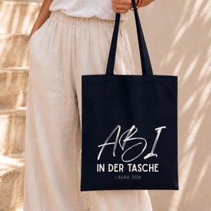 May include: A black tote bag featuring white text that reads ABI IN DER TASCHE with the name LAURA 2026 printed underneath. The bag has two long black shoulder straps and is constructed from a solid fabric material with a rectangular shape.