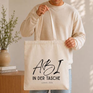 May include: A cream-colored canvas tote bag featuring black printed text that reads ABI IN DER TASCHE with the name LUKAS 2026 printed below. The bag has two long fabric handles and a rectangular shape, constructed from a light-colored textile material.