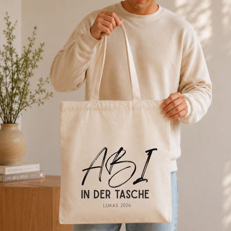 May include: A cream-colored canvas tote bag featuring black printed text that reads ABI IN DER TASCHE with the name LUKAS 2026 printed below. The bag has two long fabric handles and a rectangular shape, constructed from a light-colored textile material.