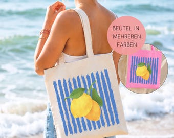 May include: A cream-colored canvas tote bag featuring a graphic print of two yellow lemons with green leaves centered over vertical blue brushstroke stripes. A circular inset shows a pink version of the same tote bag with the text BEUTEL IN MEHREREN FARBEN.