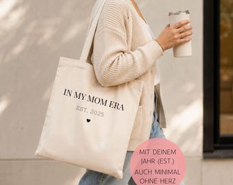 May include: A beige canvas tote bag featuring the black printed text IN MY MOM ERA, the text EST. 2025, and a small black heart symbol. The bag has two shoulder straps and is made of a light-colored fabric with a simple, rectangular design.