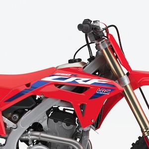 Peut inclure: Une moto tout-terrain Honda CRF 250R rouge et argentée. La moto présente une selle, des carénages et un garde-boue avant rouges. Des accents bleus et blancs sont visibles sur les carénages. Les fourches avant sont dorées. Le guidon et les commandes sont noirs.