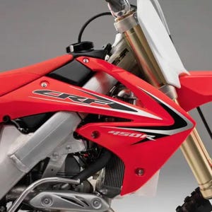 Peut inclure: Gros plan sur une moto rouge et argentée avec les inscriptions "CRF" et "450R". La moto a une selle rouge, un bouchon de réservoir noir et des fourches avant dorées. Le design est élégant et moderne.