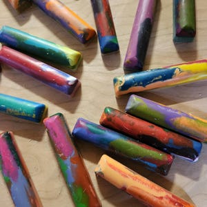 May include: A collection of colorful, rectangular crayons with a marbled appearance. The crayons are a mix of vibrant colors, including red, blue, green, and yellow, and are arranged on a light-colored wooden surface.