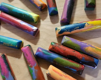 May include: A collection of colorful, rectangular crayons with a marbled appearance. The crayons are a mix of vibrant colors, including red, blue, green, and yellow, and are arranged on a light-colored wooden surface.