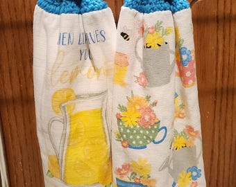 May include: Two white kitchen towels with blue crochet tops. One towel features a yellow lemonade pitcher with the text "When life gives you lemons, make lemonade." The other towel has floral and bee designs.
